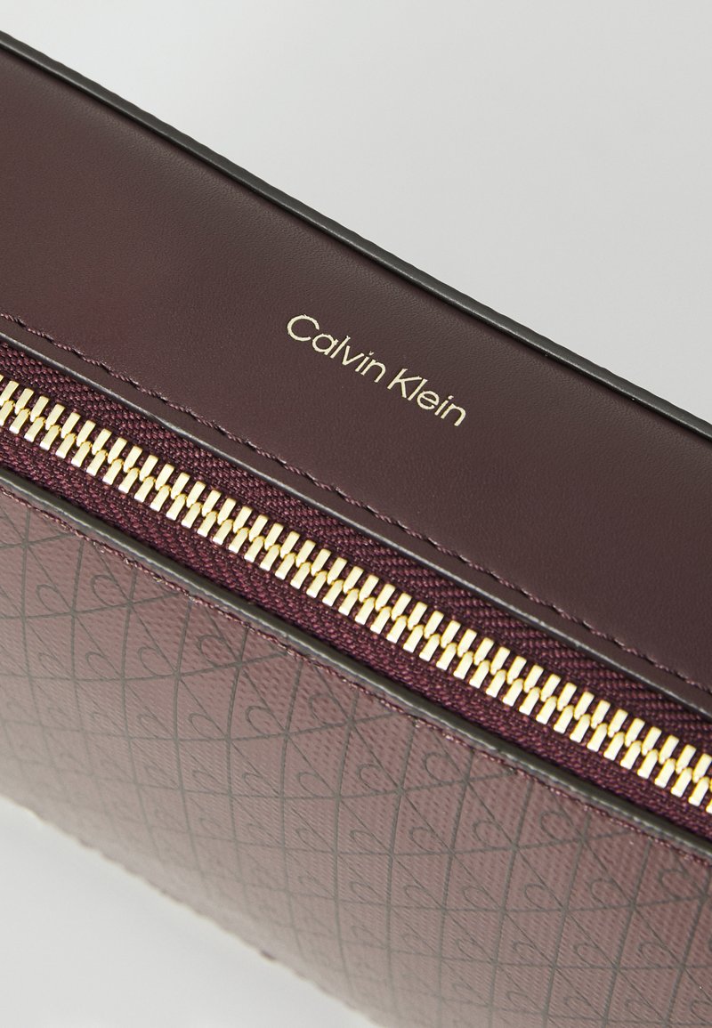 Weinrotes Calvin Klein Geldbörse mit Reißverschluss, ausgestattet mit einem strukturierten Muster und goldfarbener Reißverschlussbeschläge, mit geprägtem Logo auf der glatten Oberfläche.