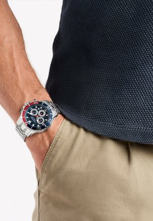 Silver metal watch with a blue dial, red accents, and three sub-dials. Worn on a wrist with a tan fabric pocket and dark textured shirt.