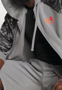 Sudadera gris con cremallera y mangas de camuflaje contrastadas, con un logo visible en naranja. Llevada sobre una camiseta negra ajustada y pantalones cortos grises.