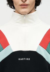 Άτομο που φοράει μπουφάν με φερμουάρ, με χρωματικό μπλοκ σε κόκκινες, τιρκουάζ, μαύρες και λευκές αποχρώσεις και το κείμενο "KAOTIKO" στο στήθος.