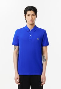 Blå polo t-shirt i slät tyg med krage, med två knappar och en grön logotyp. Kort ärm och en avslappnad passform.