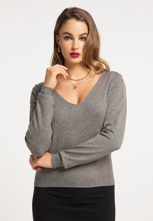 Femme aux cheveux bruns ondulés portant un pull gris à col en V, une jupe noire, des boucles d'oreilles créoles dorées, des colliers dorés superposés et du rouge à lèvres rouge, posant avec la main près du visage.