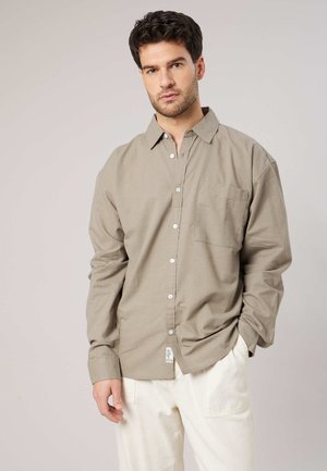 Mann mit dunklen Haaren, bekleidet mit einem beigen Hemd mit Knöpfen und weißen Hosen, steht vor einem schlichten hellen Hintergrund.