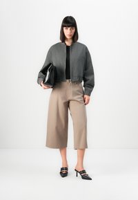 Harmaa lyhennetty bomber-takki, musta toppi, vaaleanruskeat leveälahkeiset housut, musta clutch-kotelo kädessä, mustat korot, joissa on nilkkahihnat.