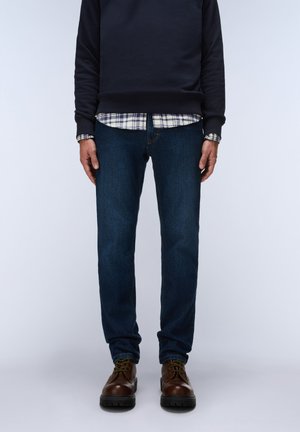 Mand iført mørkeblå jeans, brune lædersko, navy sweatshirt og en mønstret skjorte, der stikker ud under sweatshirten.