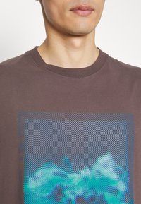 Brązowy bawełniany t-shirt z graficznym nadrukiem niebieskich i turkusowych abstrakcyjnych kształtów w kropkowanym wzorze, otoczony prostokątną ramką.