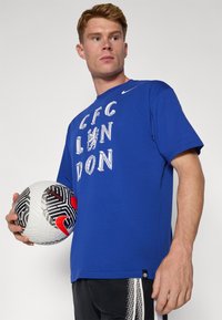 Blå bomulds T-shirt med korte ærmer, som har hvid tekst "CFC LONDON" og et løvegrafik. Modellen holder en sort, hvid og rød fodbold.