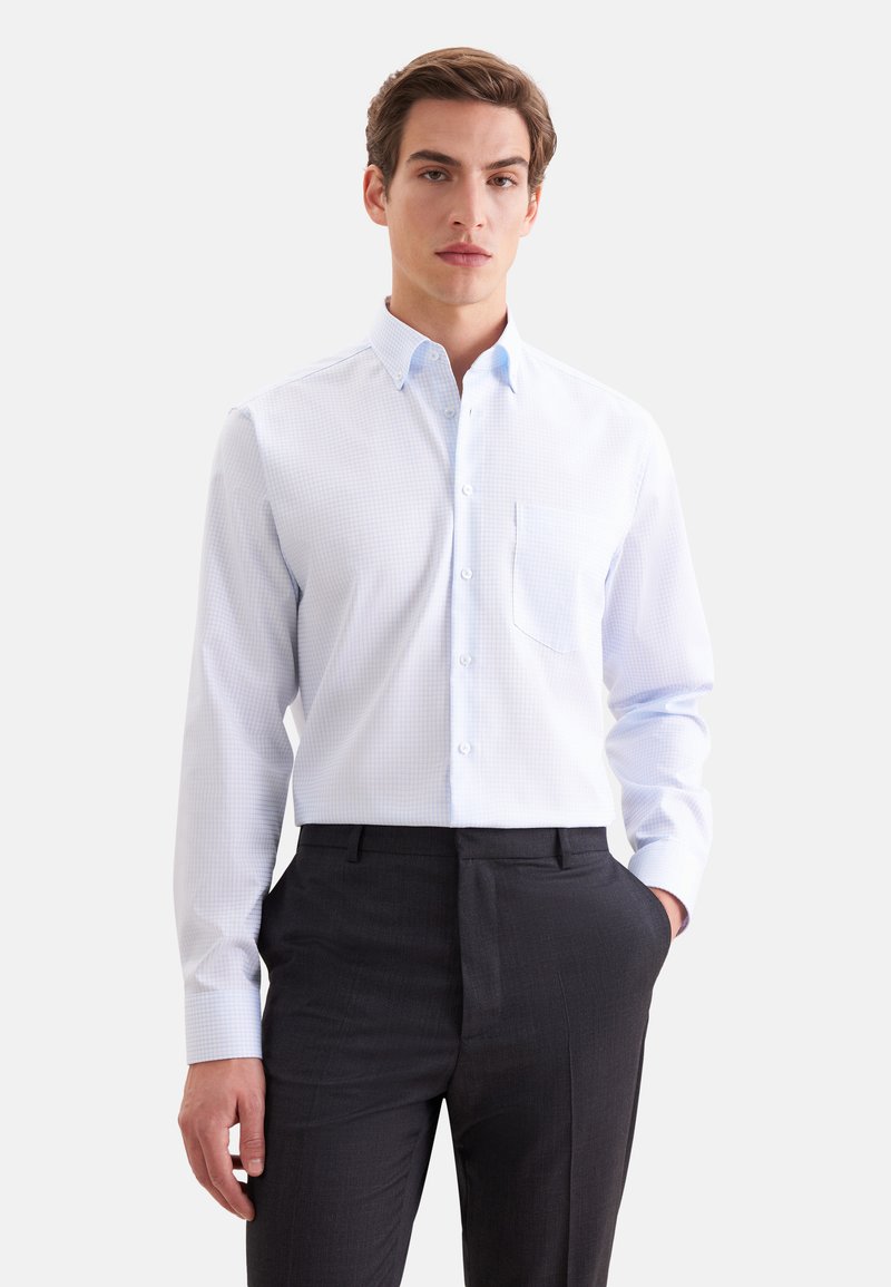 Jeune homme portant une chemise à carreaux bleu clair et un pantalon sombre, debout avec une main dans la poche devant un fond uni.