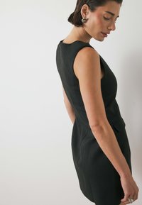Juoda suknelė, prigludusi prie kūno, be rankovių, pagaminta iš textured audinio, su maža kišenės detale. Drabužis turi švelnų kontūrą formuojantį efektą.