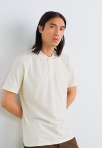 Polo in beige chiaro, maniche corte, colletto con due bottoni, tessuto liscio, branding minimale sul petto, design aderente.