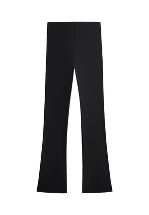 Personne portant des leggings noirs taille haute et des chaussures de sport noires, debout devant un fond gris uni.