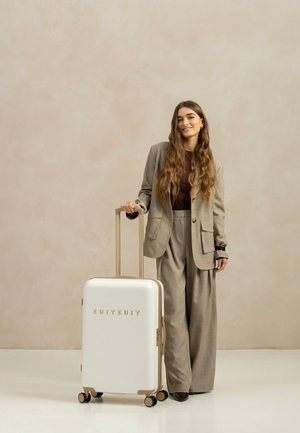 Weißer Hartschalenkoffer mit goldenen Akzenten, vier Rädern und einem teleskopischen Griff, der neben einer Person in einem beigen Anzug steht.