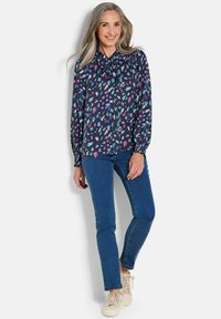 Blusa a maniche lunghe di colore blu navy con un motivo astratto colorato, colletto arricciato, abbinata a jeans blu e sneakers chiare.