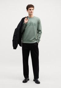 Felpa verde chiaro con logo "BOSS" in rilievo, abbinata a pantaloni neri e scarpe nere. Il modello tiene in mano una giacca nera.