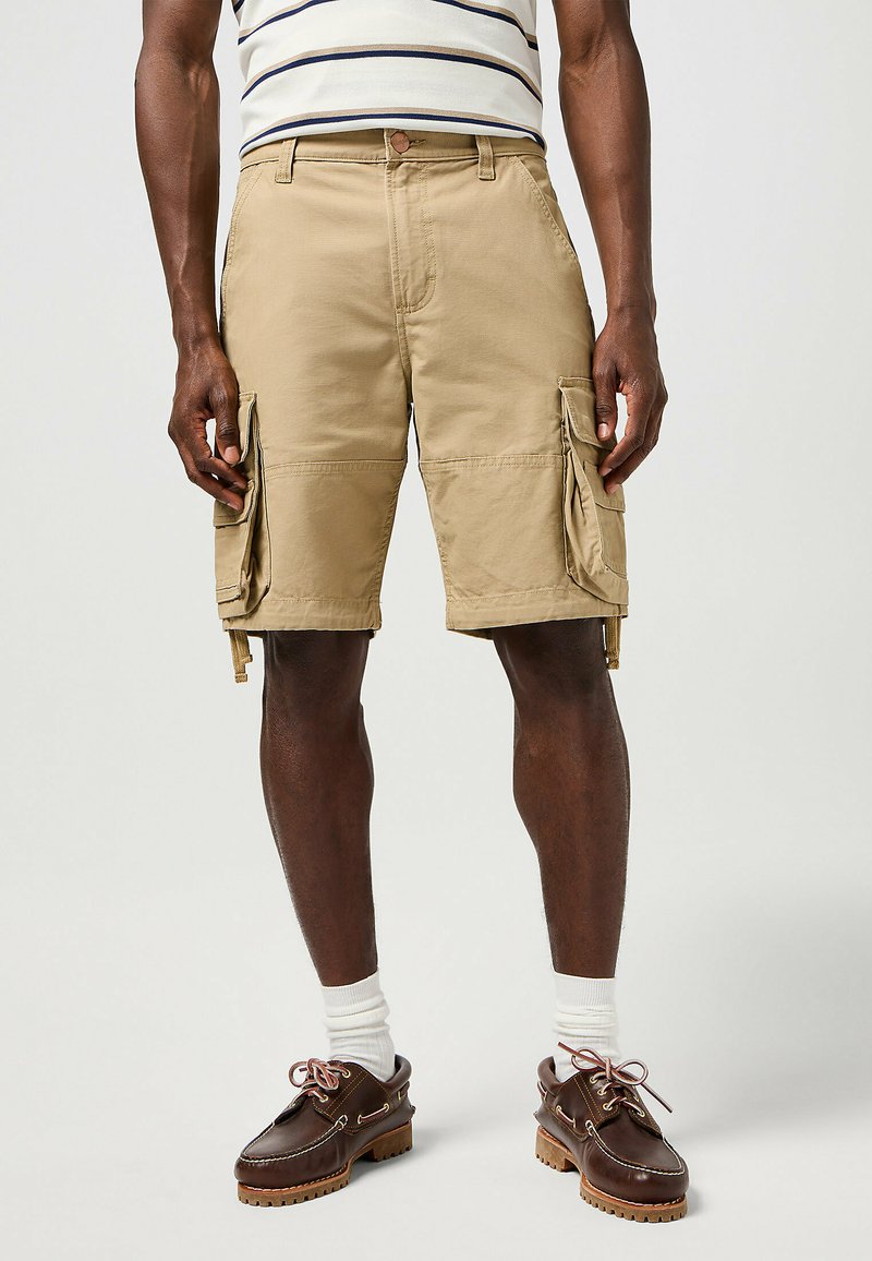 Pantaloni cargo color khaki con tasche laterali e orli regolabili, caratterizzati da una vestibilità comoda e un tessuto leggero. Indossati con scarpe marroni e calzini bianchi.