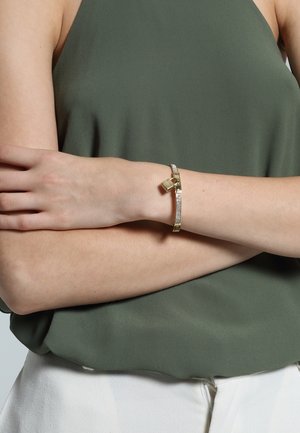 Femme portant un bracelet en or et diamants orné d'un petit pendentif en forme de cadenas, bras croisés, vêtue d'un haut sans manches vert olive et d'un pantalon blanc.