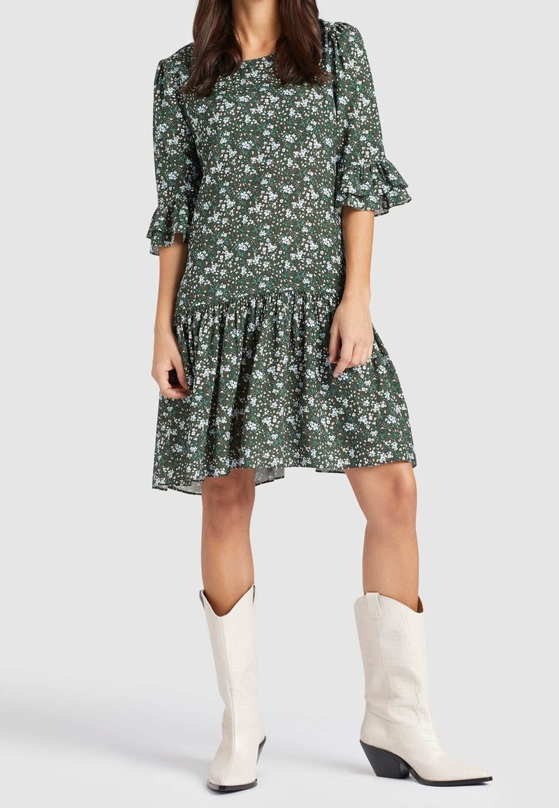 Vestido floral verde con mangas tres cuartos y detalles de volantes; a la altura de la rodilla con una falda en capas. Combinado con botas vaqueras blancas.