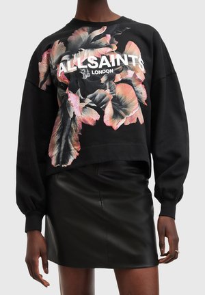 Le modèle porte un sweat-shirt noir AllSaints à imprimé floral rose et une mini-jupe en cuir noir.