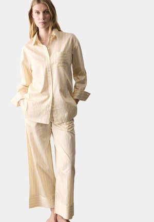 Femme blonde portant une chemise à boutons à rayures verticales jaune pâle et blanche et un pantalon large assorti avec poches, debout les mains dans les poches.