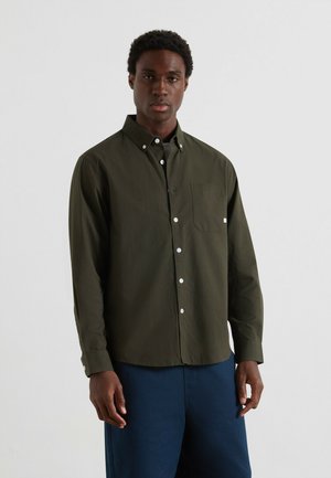 Jongeman in een donkergroene blouse met knopen en een borstzak, met een marineblauwe broek, staande tegen een effen lichte achtergrond.
