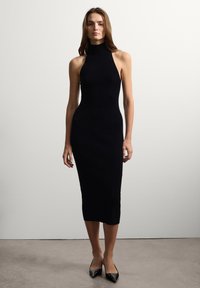 Robe midi noire à côtes avec un col montant, une silhouette ajustée et sans manches, accompagnée de talons aiguilles noirs sur un fond neutre.