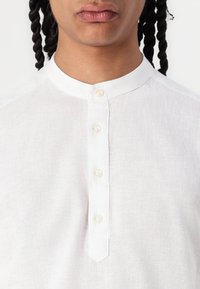 Camisa blanca de lino con cuello mandarín y un tapeta de botones con cuatro botones de perla, que muestra una tela ligera y texturada.