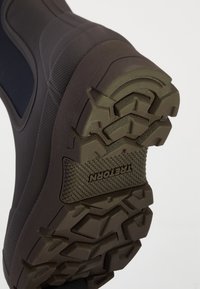 Suela de bota negra de exterior con profundas ranuras y el logo "TREKORN" grabado en la superficie de goma texturizada.