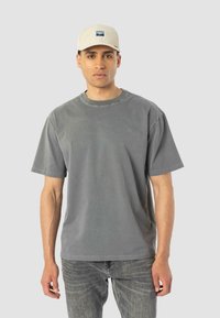 Camiseta de algodón gris con mangas cortas y cuello redondo, combinada con unos vaqueros gris ajustados y una gorra de color claro con un logo.