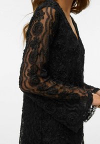 Černé krajkové šaty s květinovými ozdobami, s dlouhými průsvitnými rukávy a hlubokým výstřihem do V. Detailní textura a složitý design po celém oděvu.