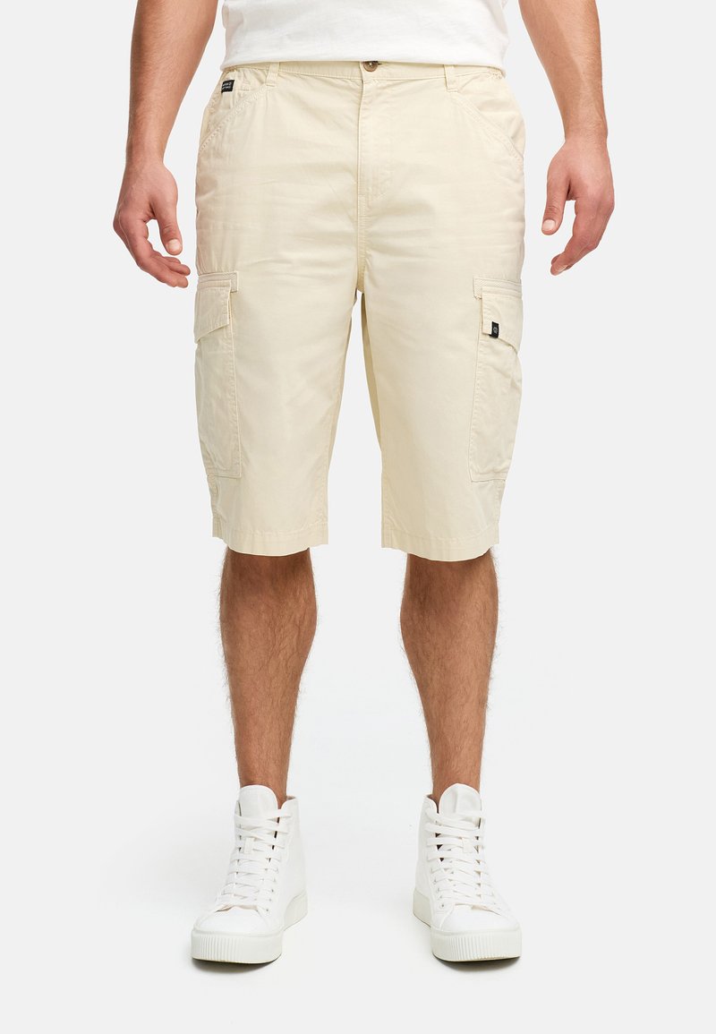 Beige cargoshorts van lichtgewicht stof met twee zijzakken, een knoopsluiting en een relaxte pasvorm, gedragen met witte high-top sneakers.