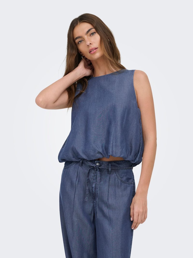 Femme aux longs cheveux bruns portant un haut en denim bleu sans manches et un pantalon taille haute assorti, posant avec une main touchant son cou.