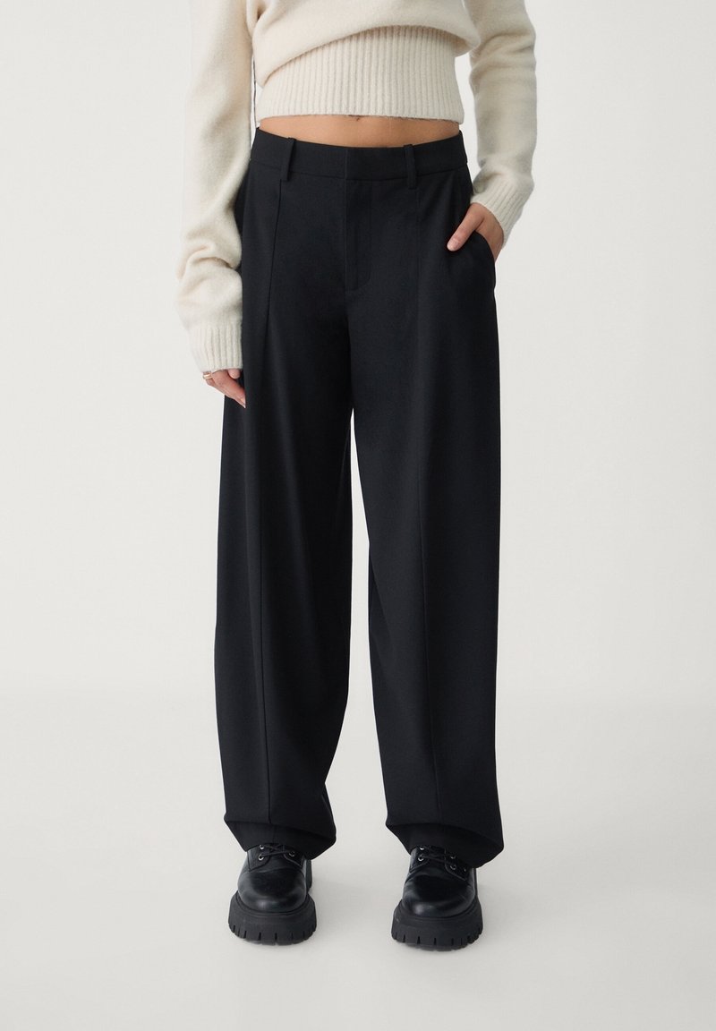 Personne portant un pantalon noir ample, des chaussures noires épaisses et un pull court crème à manches longues, debout devant un fond uni.