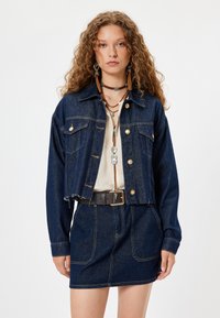 Giacca di jeans blu scuro con chiusura a bottoni dorati, orlo sfrangiato e due tasche frontali, abbinata a una gonna di jeans coordinata.