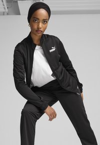 Melnā sportiskā treniņtērpa jaka un bikses ar baltu logotipu, ar rāvējslēdzēju, ribotām piedurknēm un gludu auduma tekstūru.