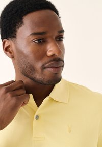 Gul polo med tekstureret stof, der har en to-knaps åbning og et lille broderet logo på venstre bryst.