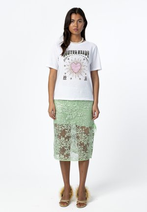 Žena v bielom tričku s potlačou ružového srdca a zelenou midi sukňou s čipkou, doplnená perlovým náhrdelníkom a sandálmi s kožušinovým lemom.