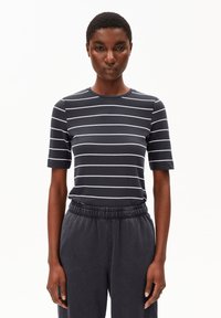 Vrouw in een zwart-witte horizontaal gestreepte T-shirt met korte mouwen en zwarte elastische broeken staat tegen een witte achtergrond.