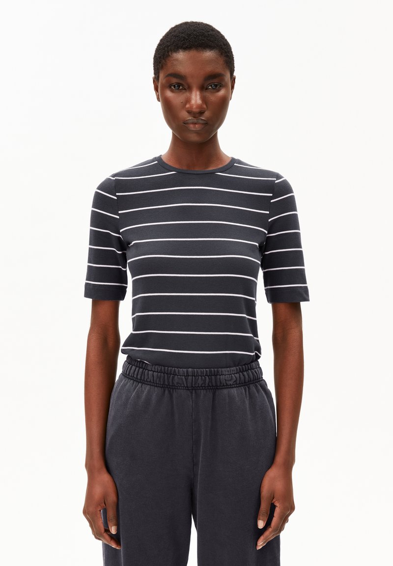 Vrouw in een zwart-witte horizontaal gestreepte T-shirt met korte mouwen en zwarte elastische broeken staat tegen een witte achtergrond.