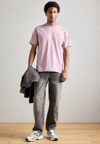 Ανοιχτό ροζ βαμβακερό t-shirt με κοντά μανίκια, συνδυασμένο με γκρι τζιν παντελόνι και λευκά sneakers με μαύρες λεπτομέρειες. Κρατάει μία γκρίζα ζακέτα.