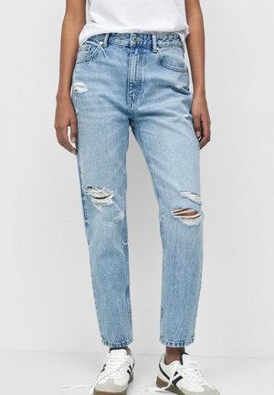 Personne portant un jean bleu clair déchiré, des baskets blanches avec des bandes noires, et un t-shirt blanc sur un fond clair uni.