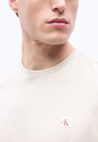 Νεαρός άνδρας που φοράει ανοιχτό μπεζ μπλουζάκι με στρογγυλή λαιμόκοψη και μικρό κόκκινο και λευκό έμβλημα λογοτύπου Calvin Klein στο στήθος.