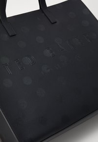 Sac fourre-tout noir texturé avec des pois en relief, doté de poignées plates et du nom de la marque "TED BAKER LONDON" affiché en évidence.