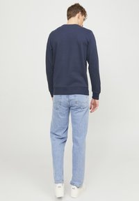 Marineblauwe katoenen sweatshirt met een ronde halslijn en ribgebreide mouwen, gedragen met lichtblauwe jeans en witte sneakers, gezien vanuit de rug.