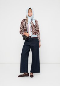 Braun gemusterte Strickjacke mit Knöpfen, blauen Akzenten, weite dunkelblaue Jeans und glänzende burgunderfarbene Ballerinas. Blau gemusterter Kopftuch.