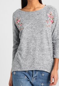 Maglione grigio texture con maniche a tre quarti, caratterizzato da ricami floreali rosa e marroni sul petto. Abbinato a jeans decorati.