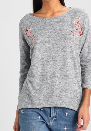 Maglione grigio texture con maniche a tre quarti, caratterizzato da ricami floreali rosa e marroni sul petto. Abbinato a jeans decorati.