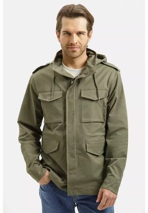 Mężczyzna w oliwkowo zielonej kurtce z kapturem, z przednimi kieszeniami i zamkiem błyskawicznym, w połączeniu z niebieskimi dżinsami, na jednolitym białym tle.