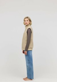 Beige Fleece-Weste mit hohem Kragen, Reißverschlusstaschen und ärmellosem Design, getragen über einem braunen Langarmoberteil und kombiniert mit blauen Jeans.
