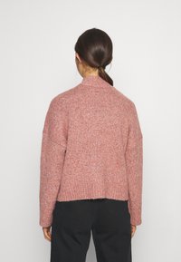 Pull en maille rose texturée avec un col montant, des épaules tombantes et un ourlet côtelé, associé à un pantalon noir taille haute.