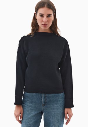 Maglione nero lavorato a maglia con scollo tondo, maniche lunghe e dettagli in oro sui bottoni sulle spalle. Il materiale sembra morbido e strutturato.